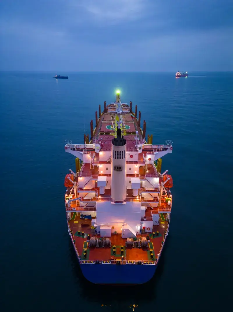 A loaded bulk carrier lying at anchor at dusk, seen bow-on from the air, with other vessels on the horizon
